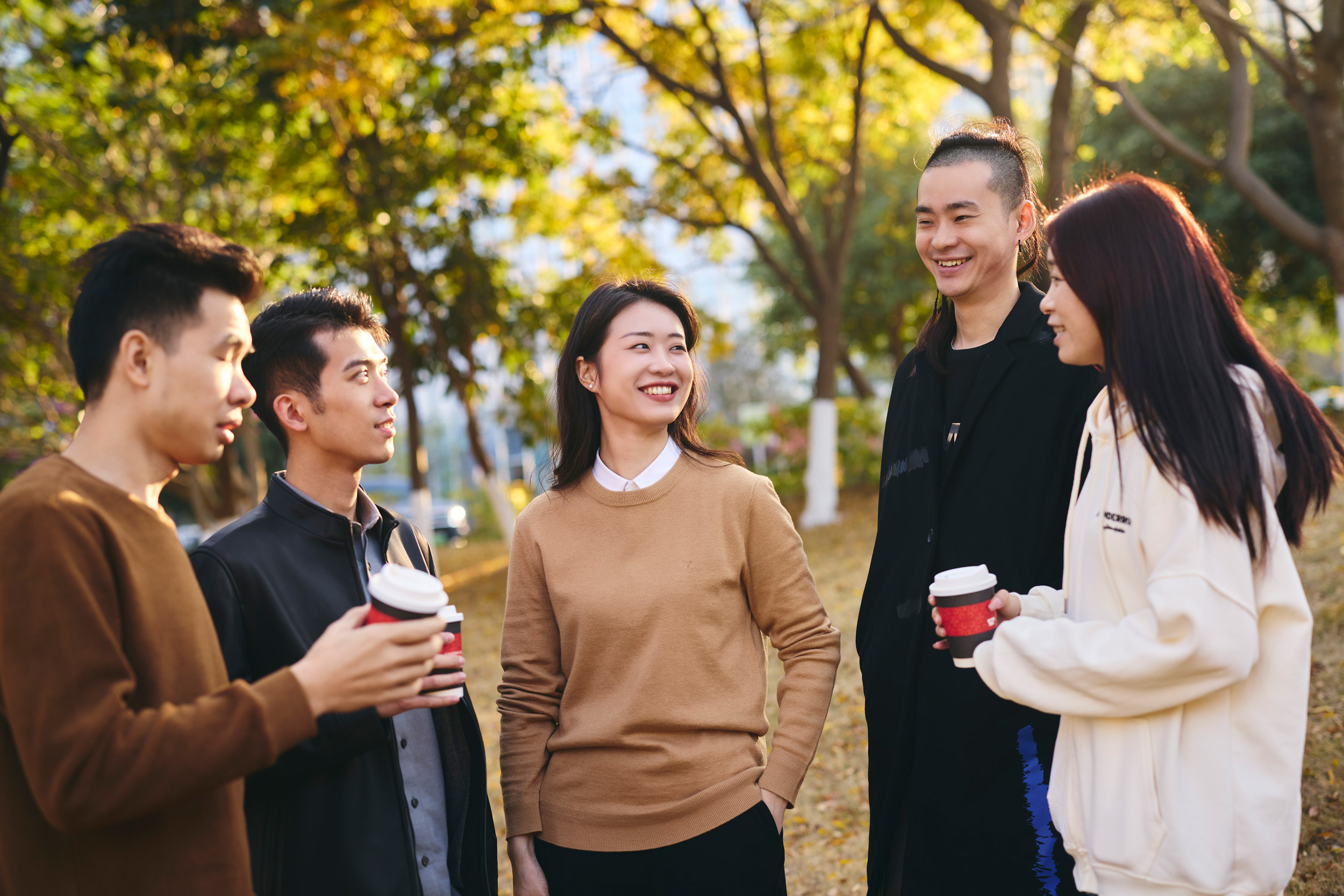 Group of young adults chatting outdoors with coffee cups in a park setting.